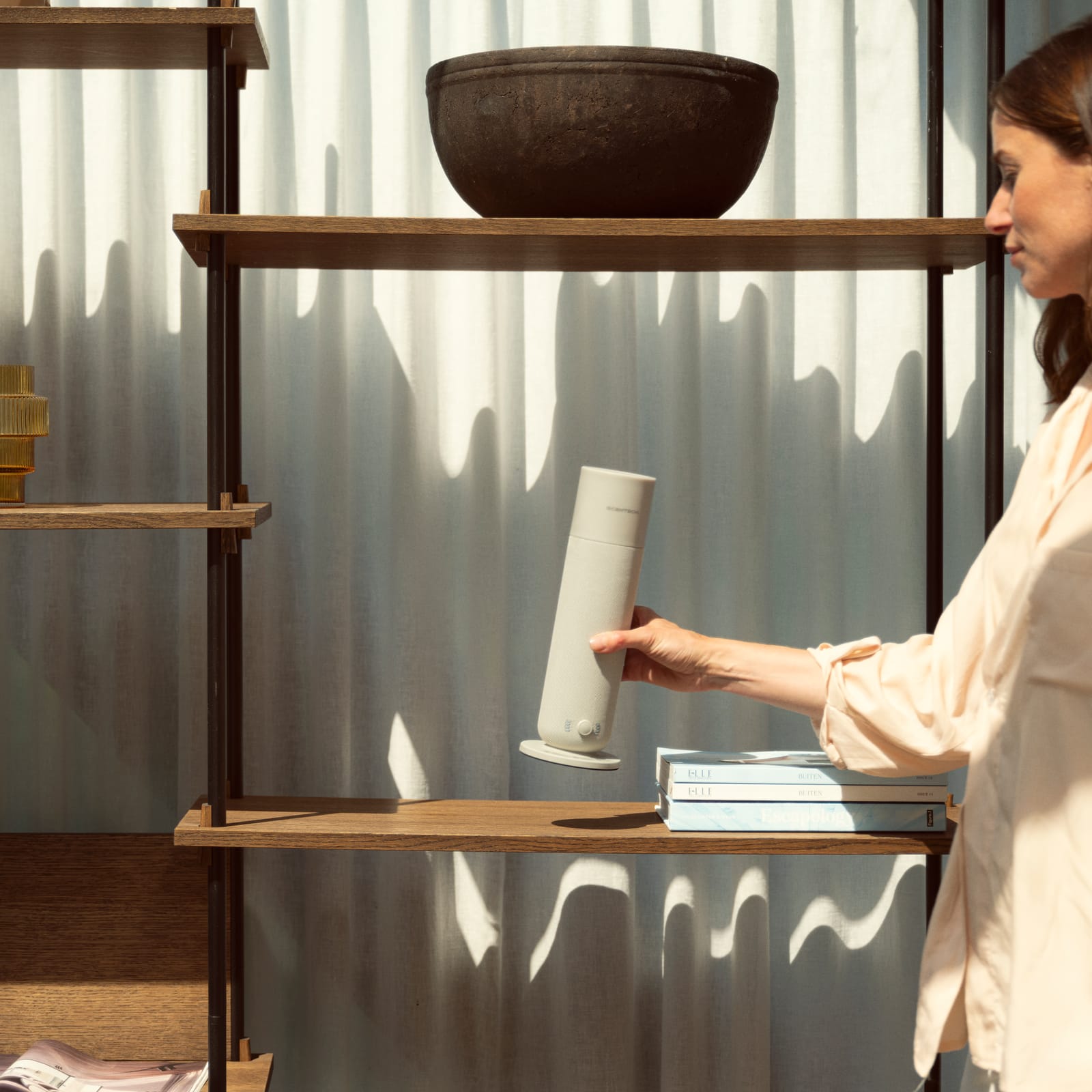 Eine Frau stellt den Scentech Smart Aroma Diffuser 2 auf ein Holzregal vor dem Vorhang